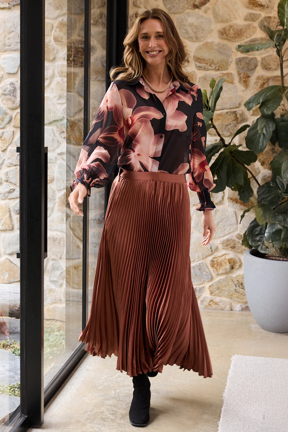 Woman wearing a floral blouse and pleated skirt standing indoors with a stone wall and plant in the background.