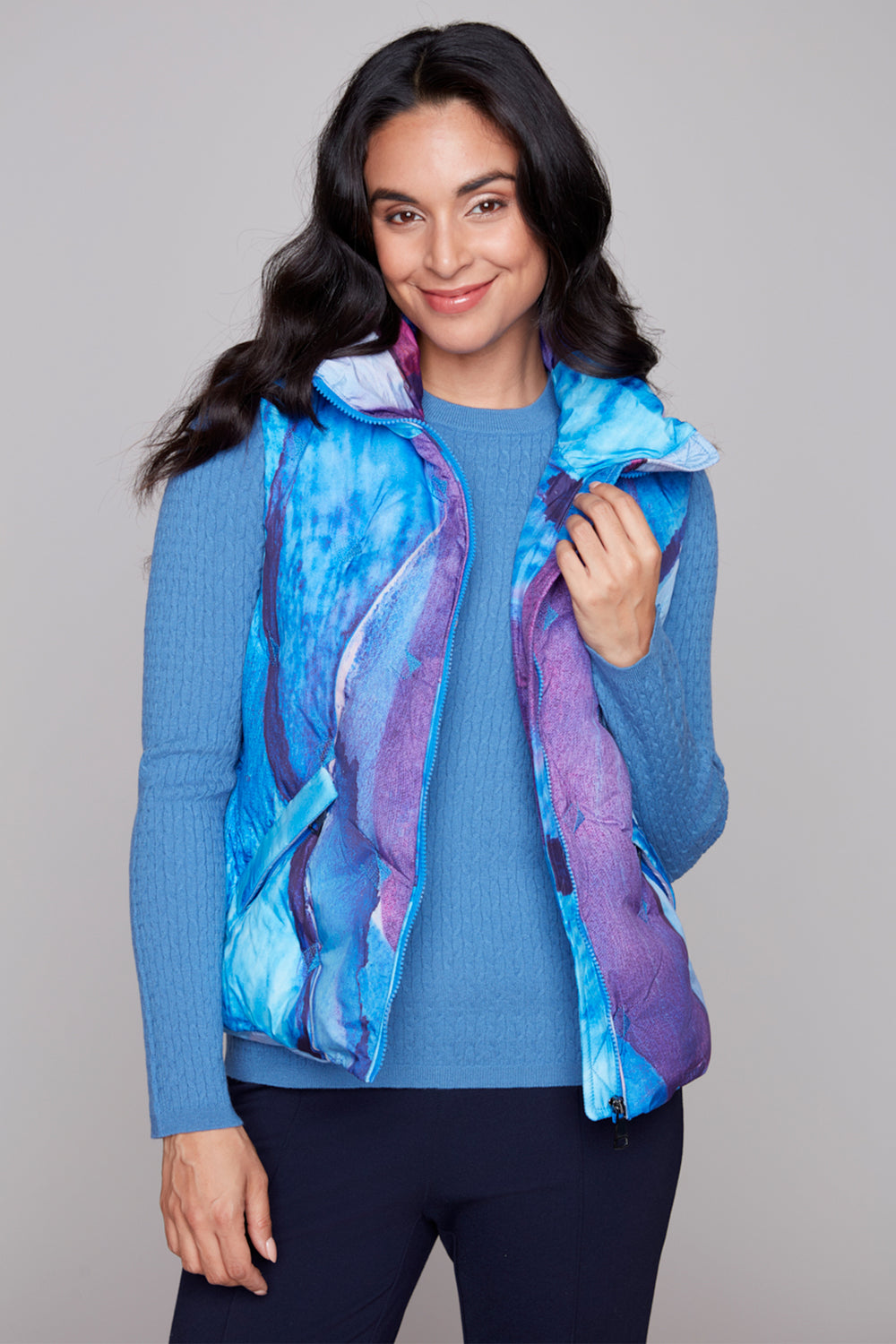 Woman wearing a blue and purple patterned vest over a blue sweater on a gray background