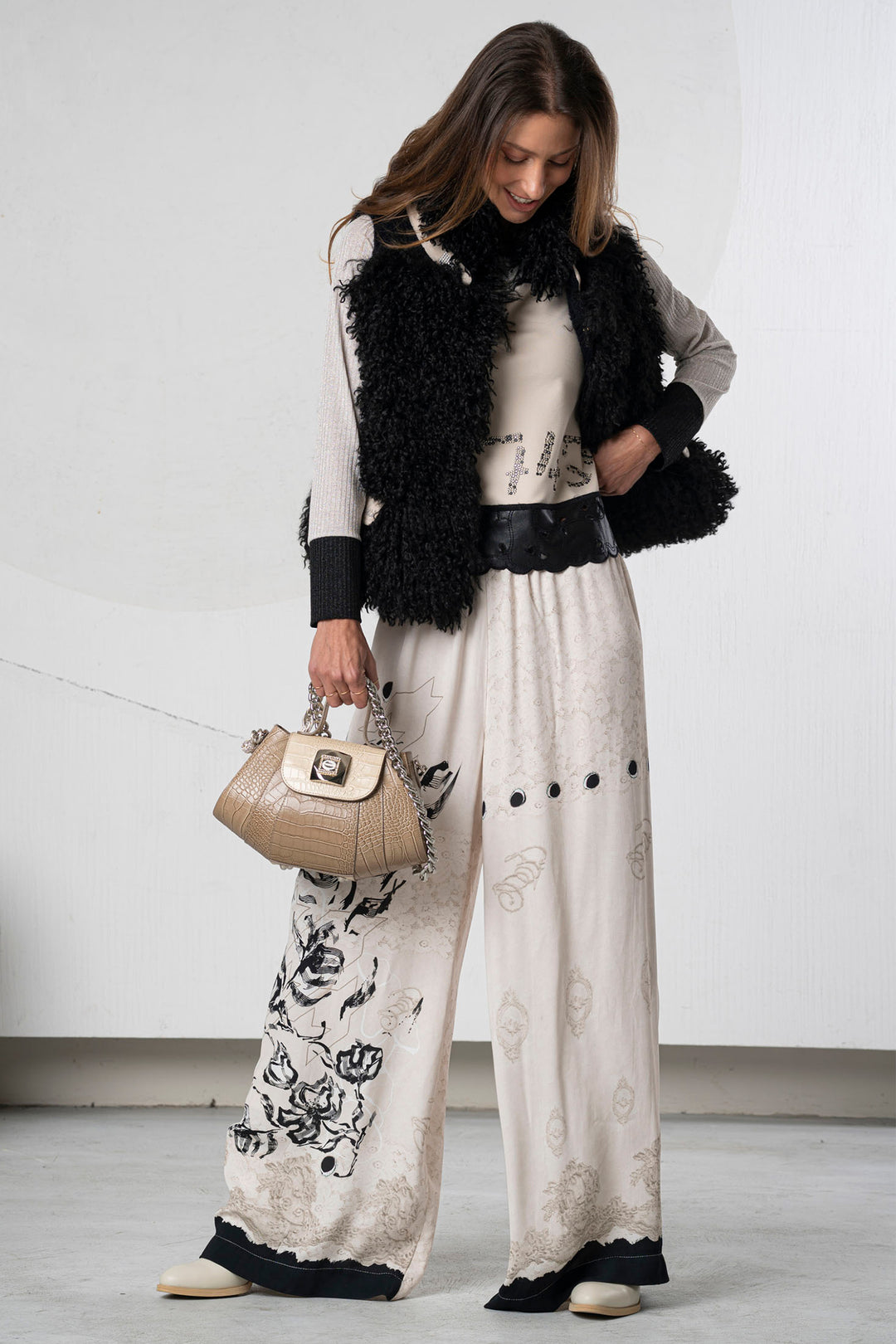 Woman wearing a stylish outfit with a black fur vest, white top, and patterned pants against a plain background.