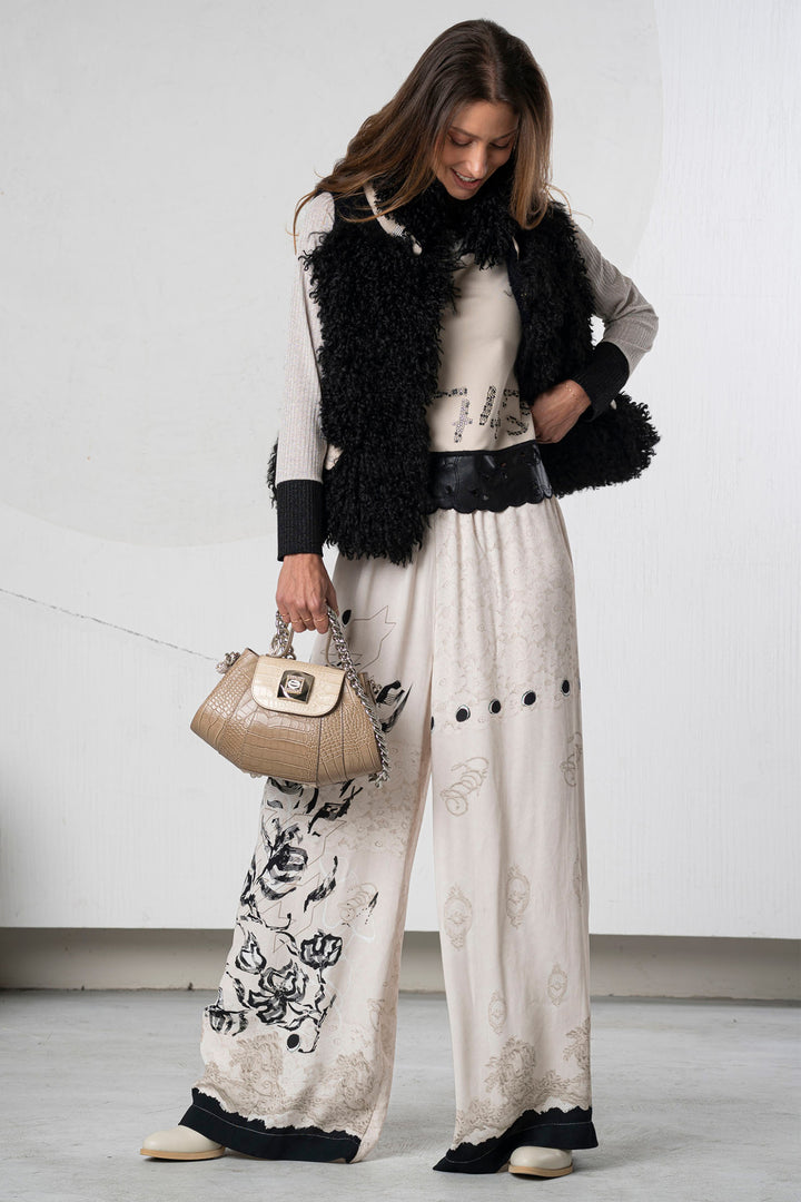 Woman wearing a stylish outfit with a black fur vest, white top, and patterned pants against a plain background.