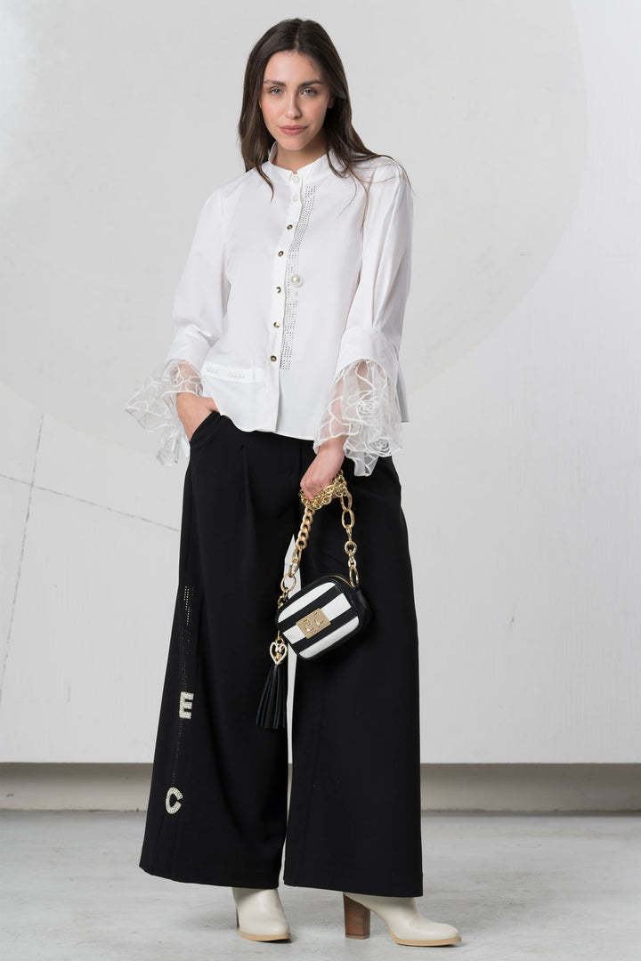 Woman wearing a white blouse with lace sleeves and black pants, holding a striped handbag.