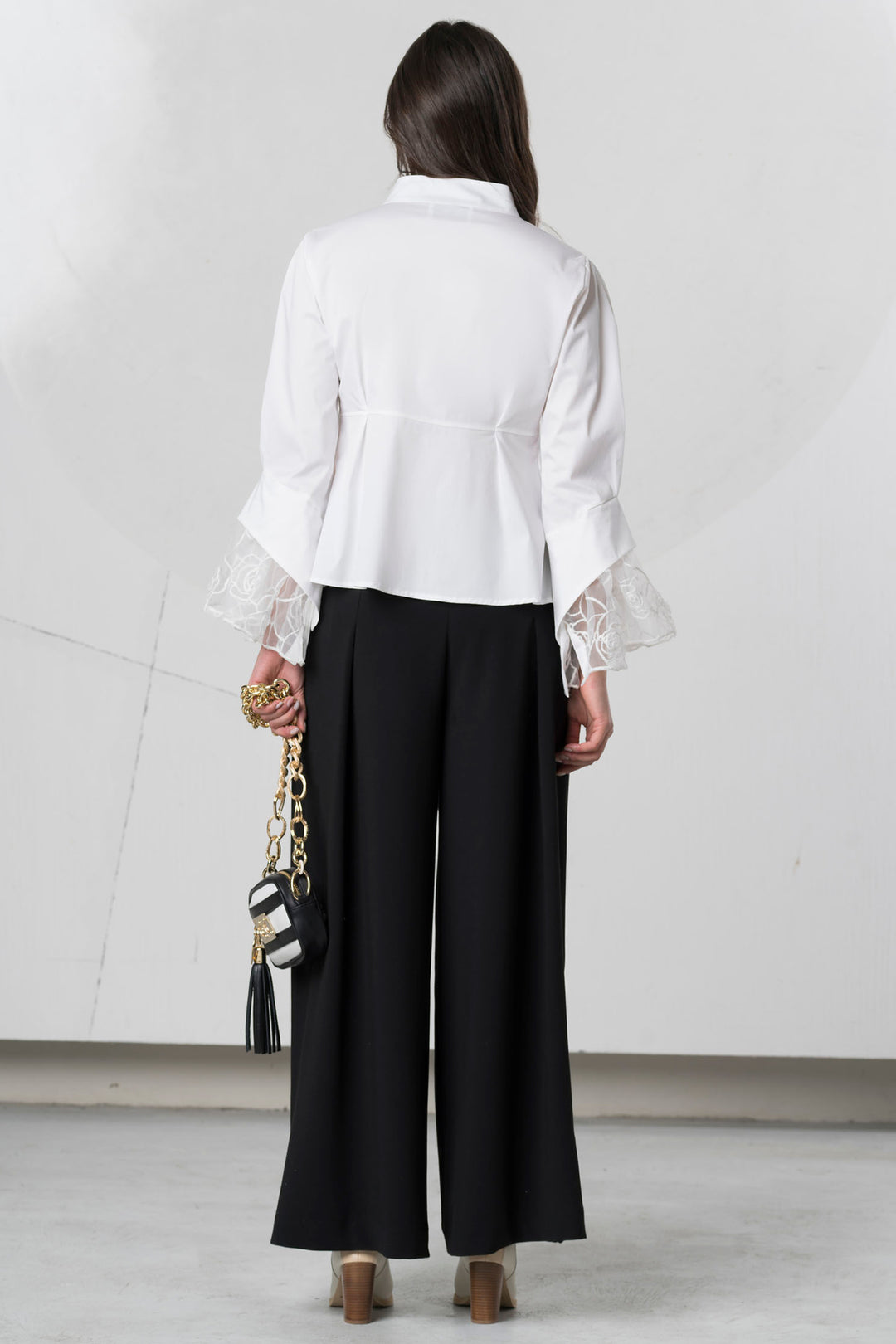 Person wearing a white blouse with ruffled sleeves and black pants, holding a black handbag.