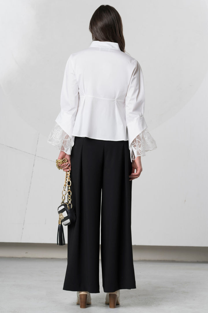 Person wearing a white blouse with ruffled sleeves and black pants, holding a black handbag.