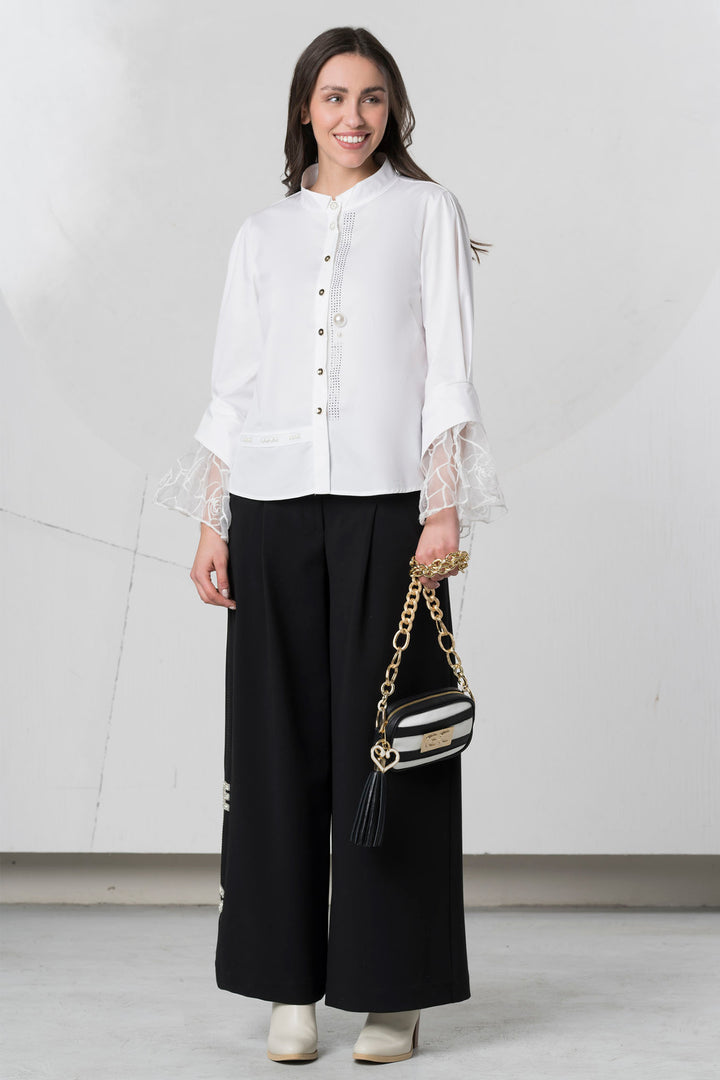 Woman wearing a white blouse with sheer sleeves and black pants, holding a handbag against a white background.