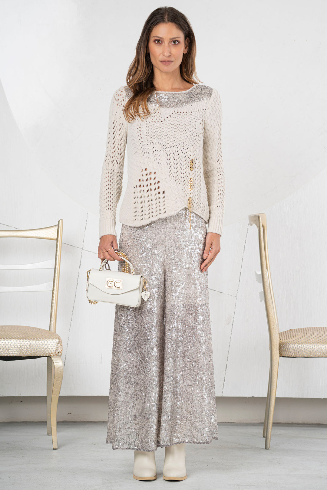 Woman wearing a white lace top and silver sequin pants, holding a white handbag, standing in a minimalistic room with white walls and gold chairs.