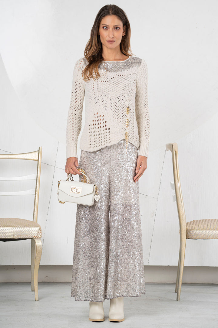 Woman wearing a white lace top and silver sequin pants, holding a white handbag, standing in a minimalistic room with white walls and gold chairs.