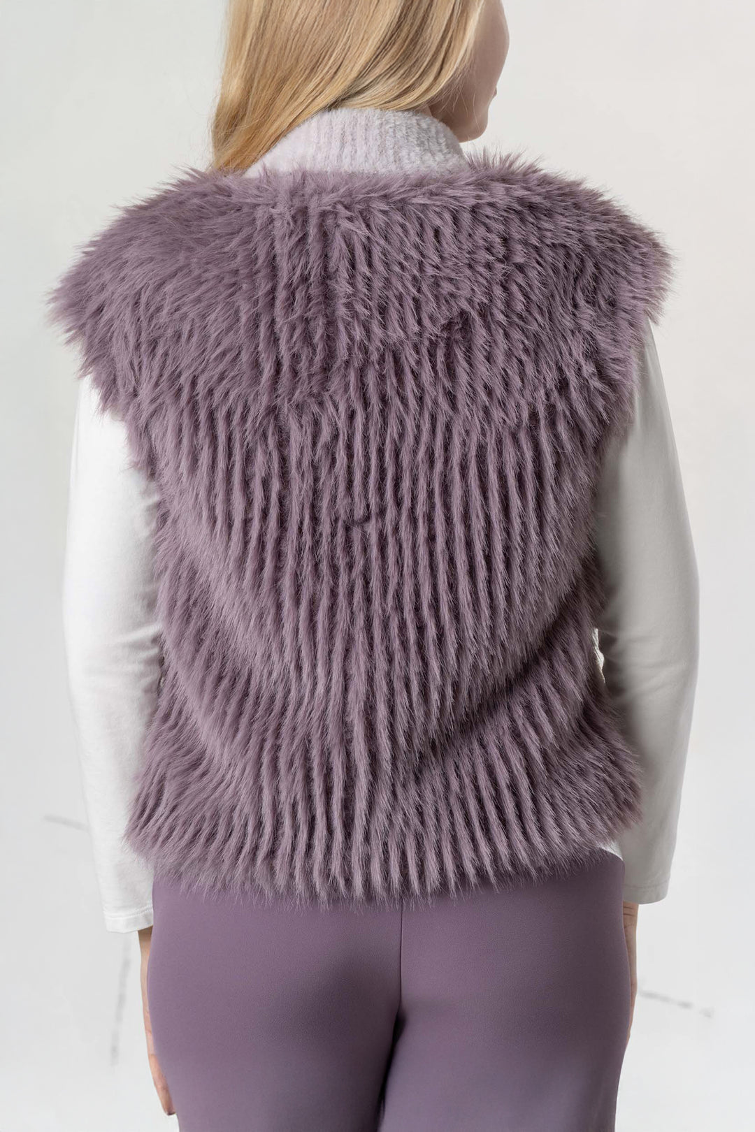 Person wearing a fluffy purple vest over a white shirt and purple pants on a light gray background