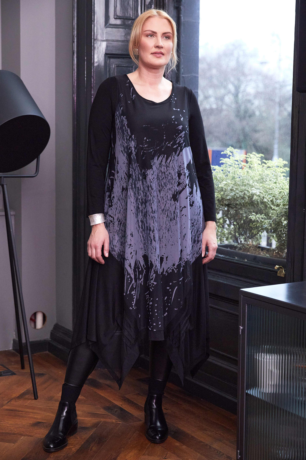 Woman wearing a black and gray dress standing indoors with a window in the background.