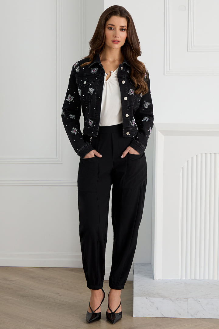 Woman wearing a black floral embroidered jacket and black pants against a white paneled wall.