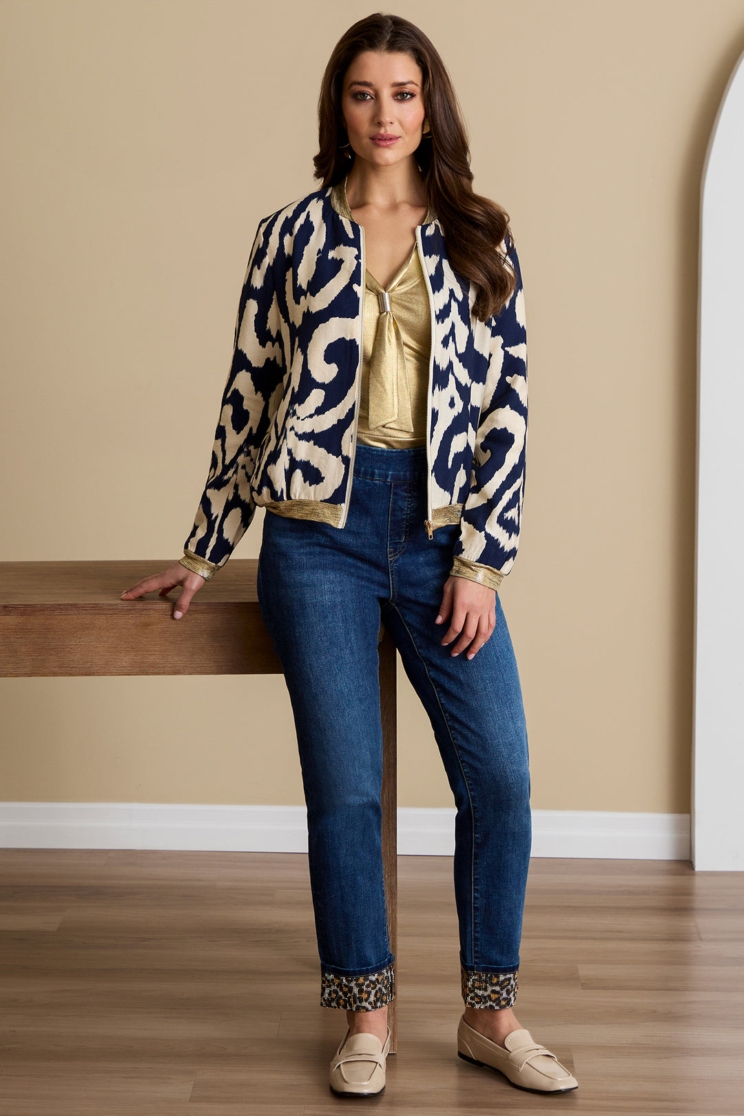 Woman wearing a patterned jacket, gold blouse, and blue jeans against a beige wall.