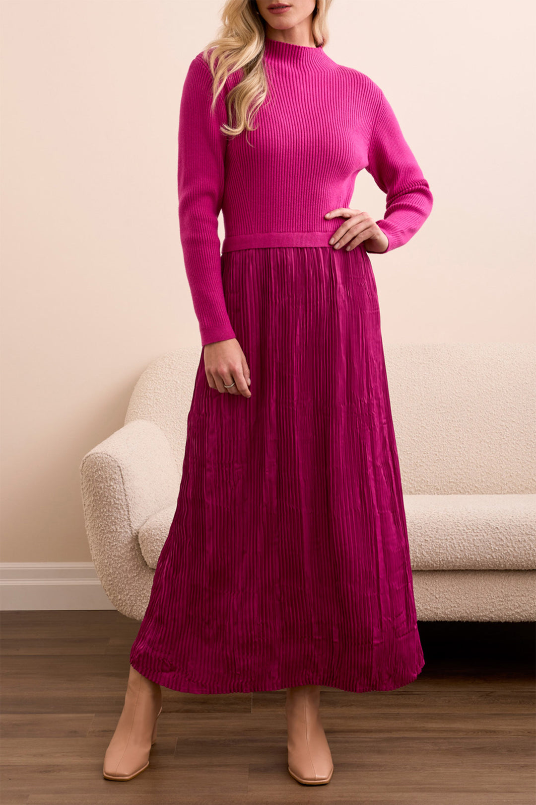 Woman wearing a pink ribbed sweater and pleated skirt standing in a room with a beige sofa.