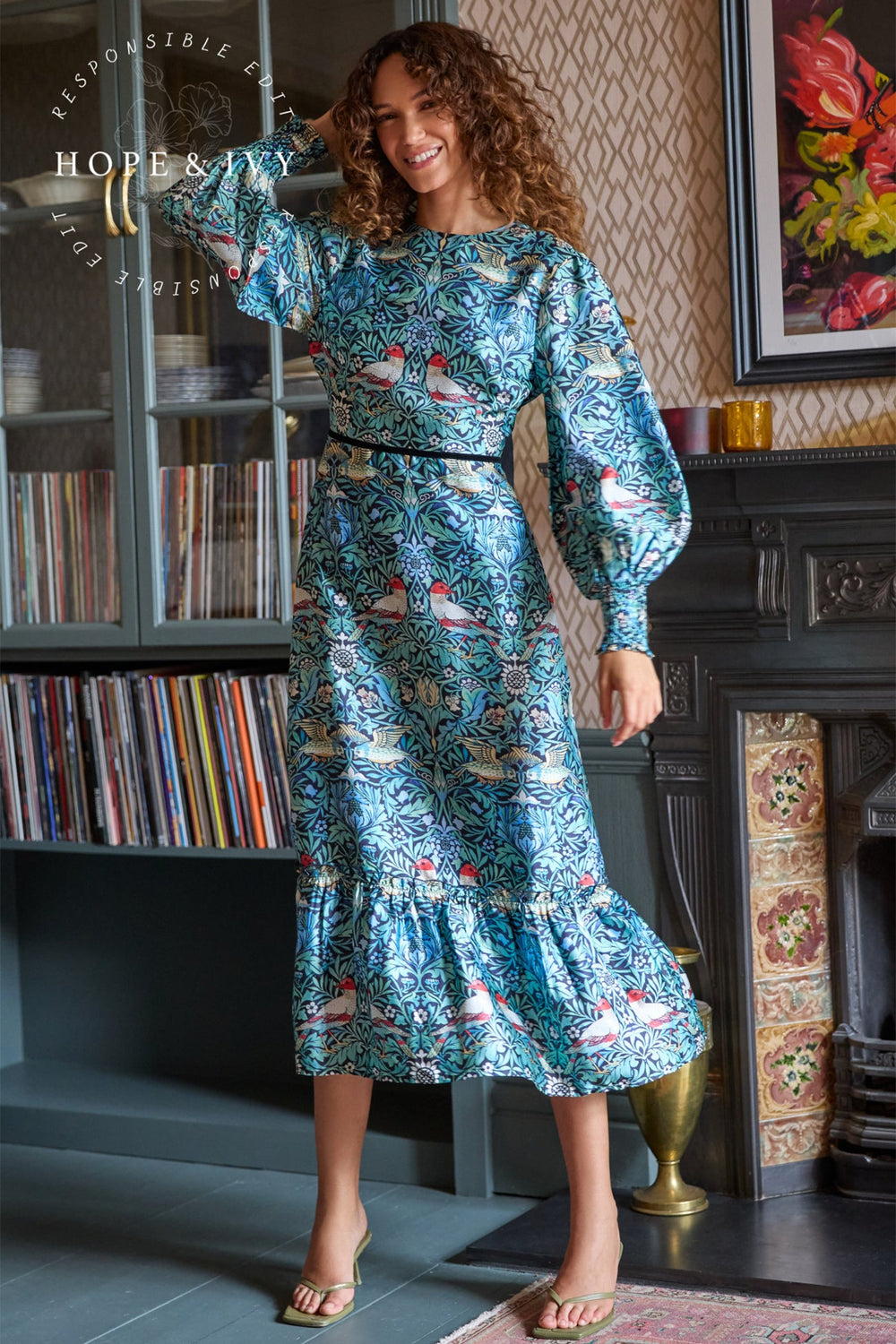 Woman in a blue patterned dress standing in a room with a fireplace and bookshelf.
