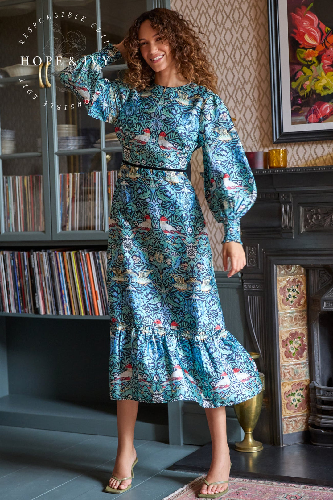 Woman in a blue patterned dress standing in a room with a fireplace and bookshelf.