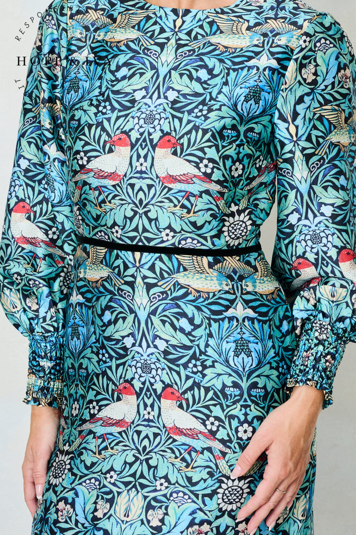 Woman wearing a dress with a detailed pattern of birds and foliage on a teal background.