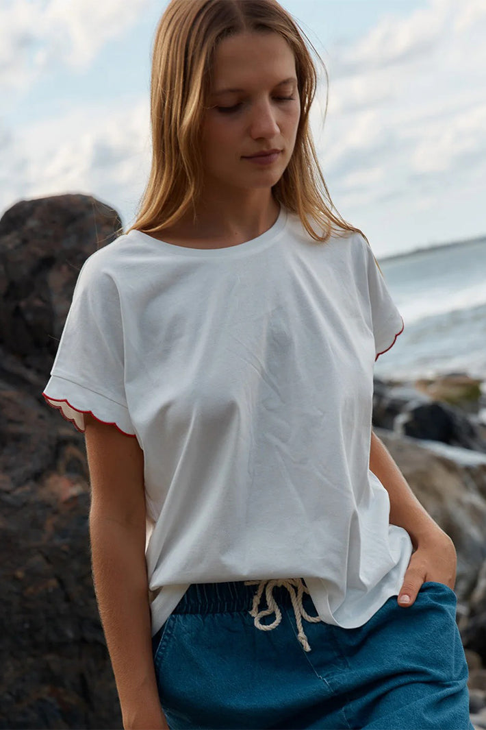 Woman wearing a white t-shirt with red trim and blue pants standing on a rocky beach.