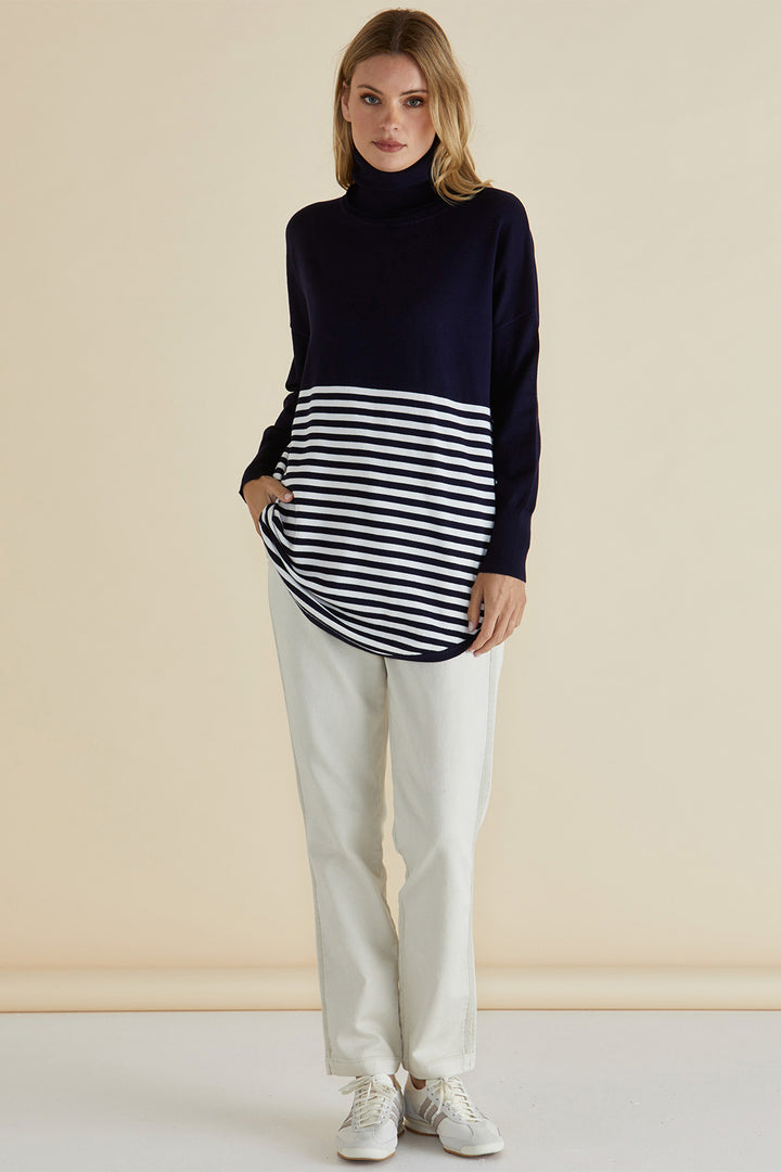 Woman wearing a navy turtleneck sweater over a black and white striped top with light-colored pants against a beige background.