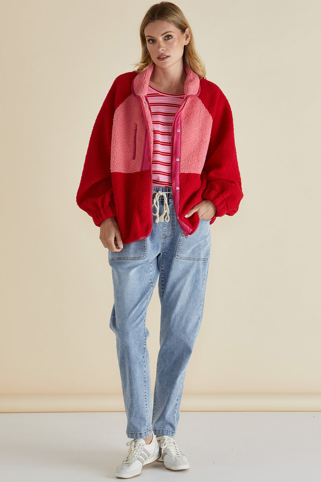 Woman wearing a red jacket, striped shirt, and blue jeans against a beige background