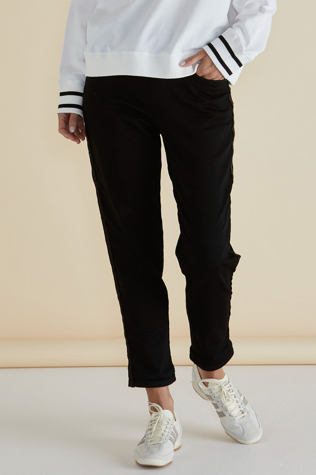 Person wearing a white sweatshirt with black and white striped cuffs, black pants, and white sneakers on a beige background