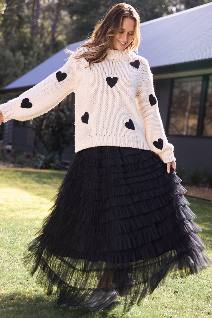 Woman wearing a white sweater with black heart patterns and a black fringed skirt outdoors.