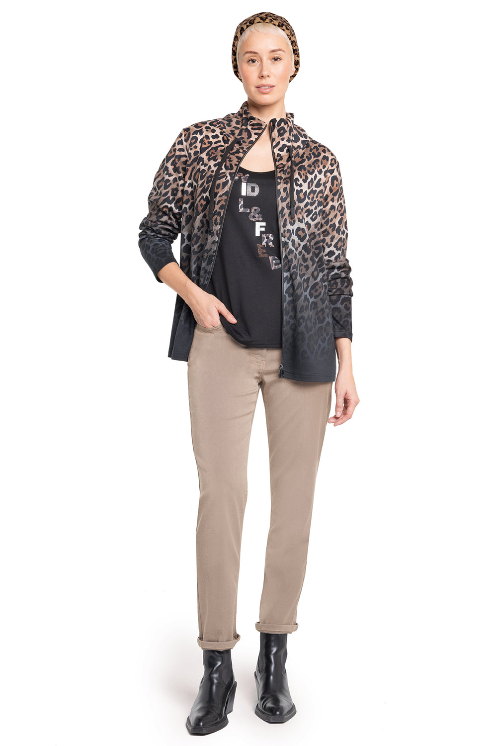 Woman wearing a leopard print shirt over a black top with text, beige pants, and black boots on a white background