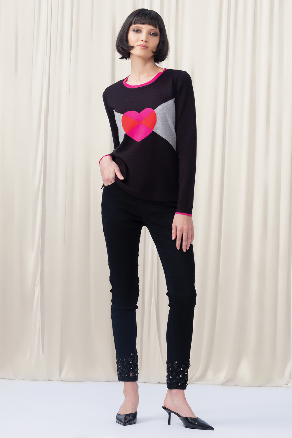 Woman wearing a black sweater with a colorful heart design and black pants against a white curtain background