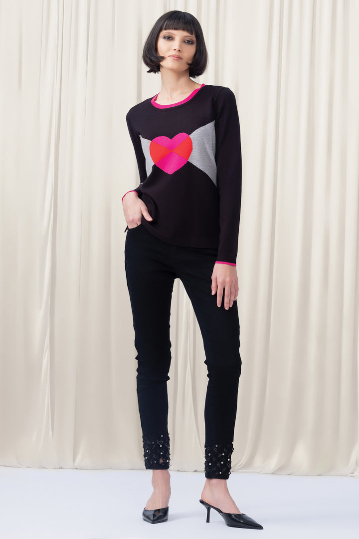 Woman wearing a black sweater with a colorful heart design and black pants against a white curtain background