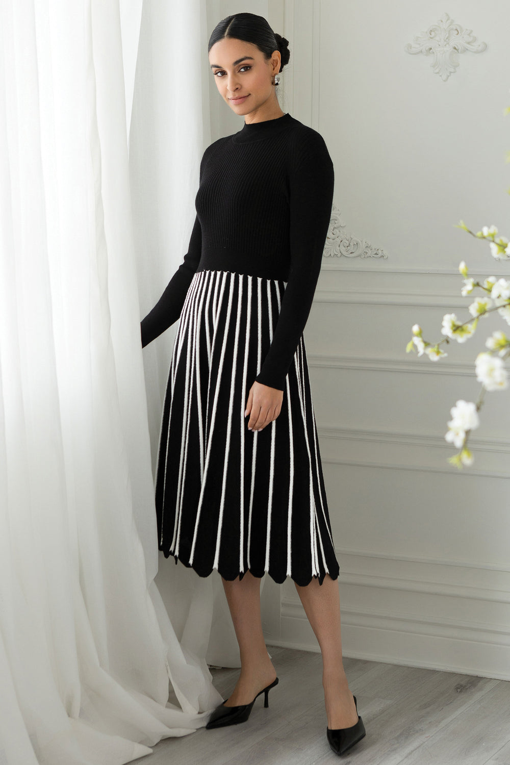 Woman wearing a black top and black and white striped skirt standing in a room with white curtains and a light-colored wall.