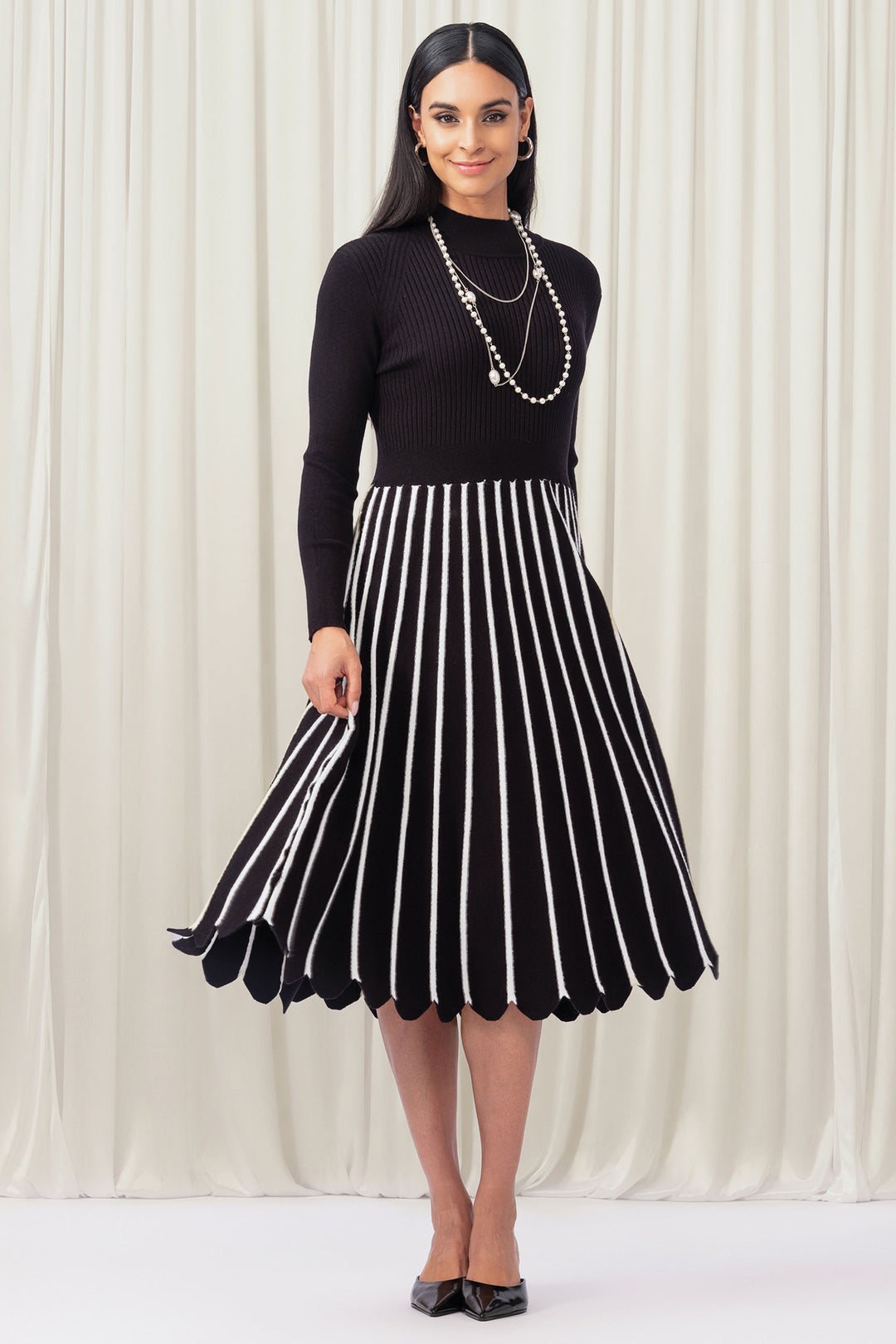Woman wearing a black top and black and white striped skirt against a white curtain background