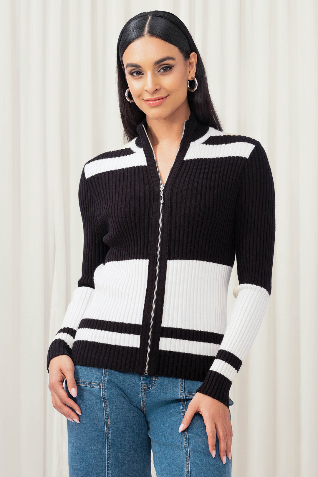 Woman wearing a black and white striped sweater with a zipper, standing against a white curtain background.