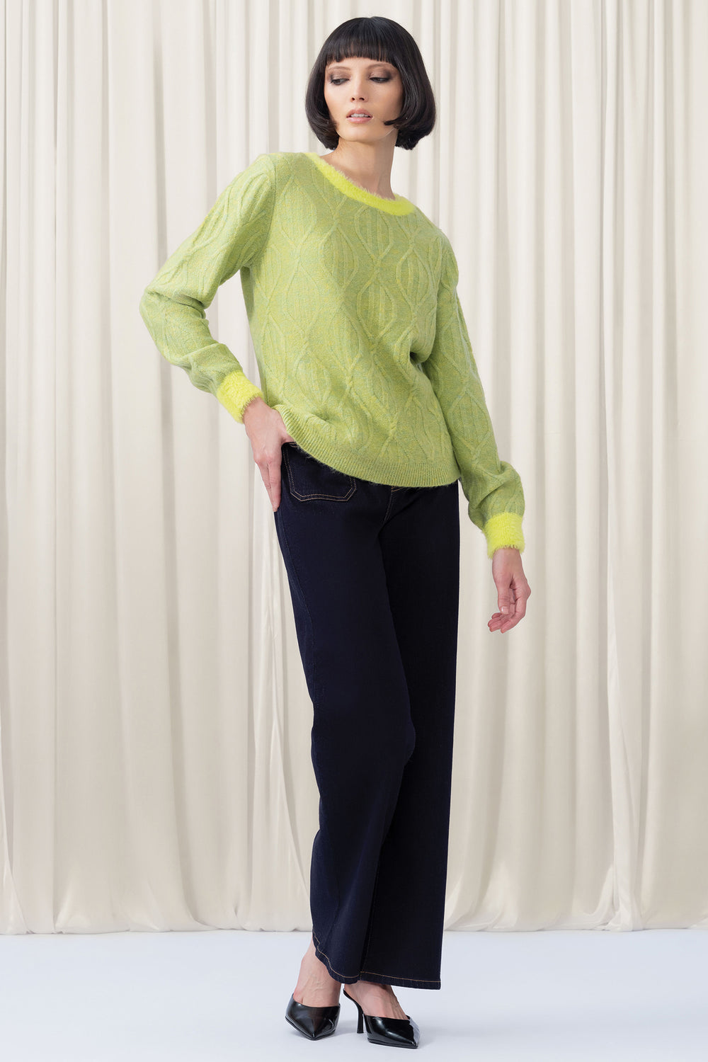 Woman wearing a green sweater and dark pants against a white curtain background