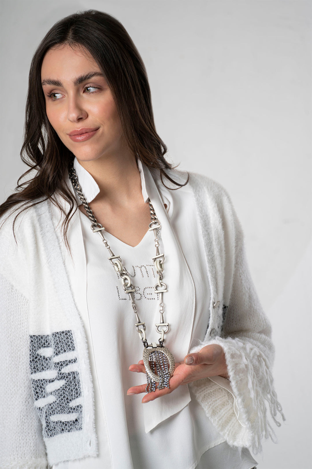 Woman wearing a white scarf with fringe, holding a large silver necklace against a plain background