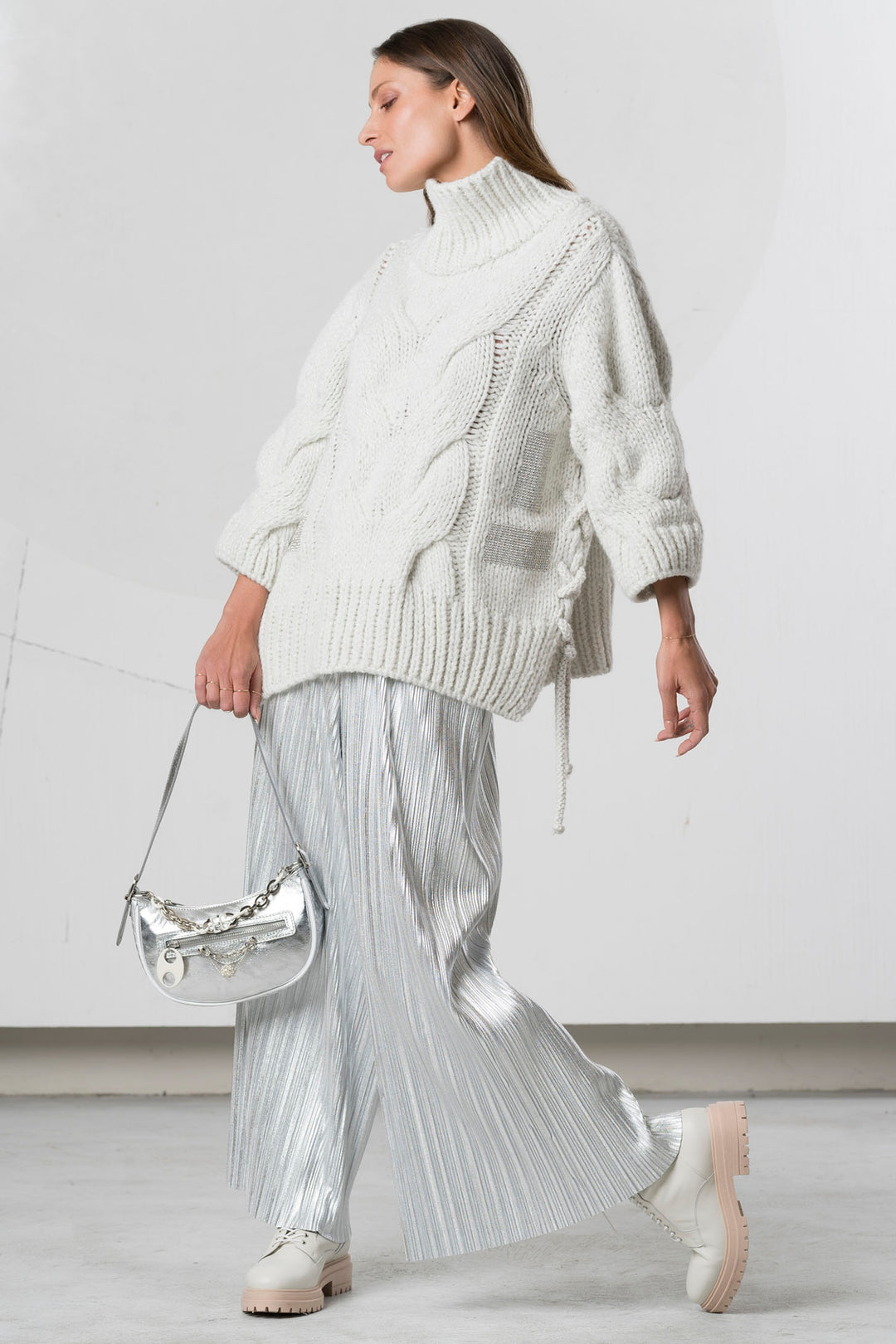 Woman wearing a white knit sweater and silver pleated skirt against a plain background