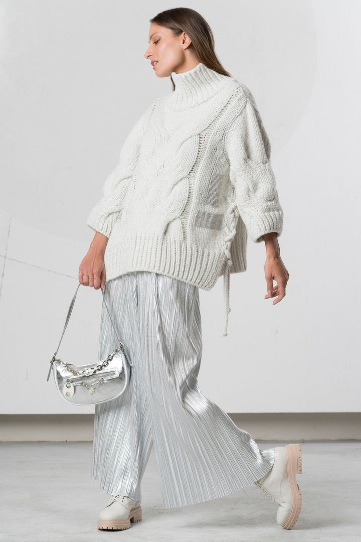 Woman wearing a white knit sweater and silver pleated skirt against a plain background