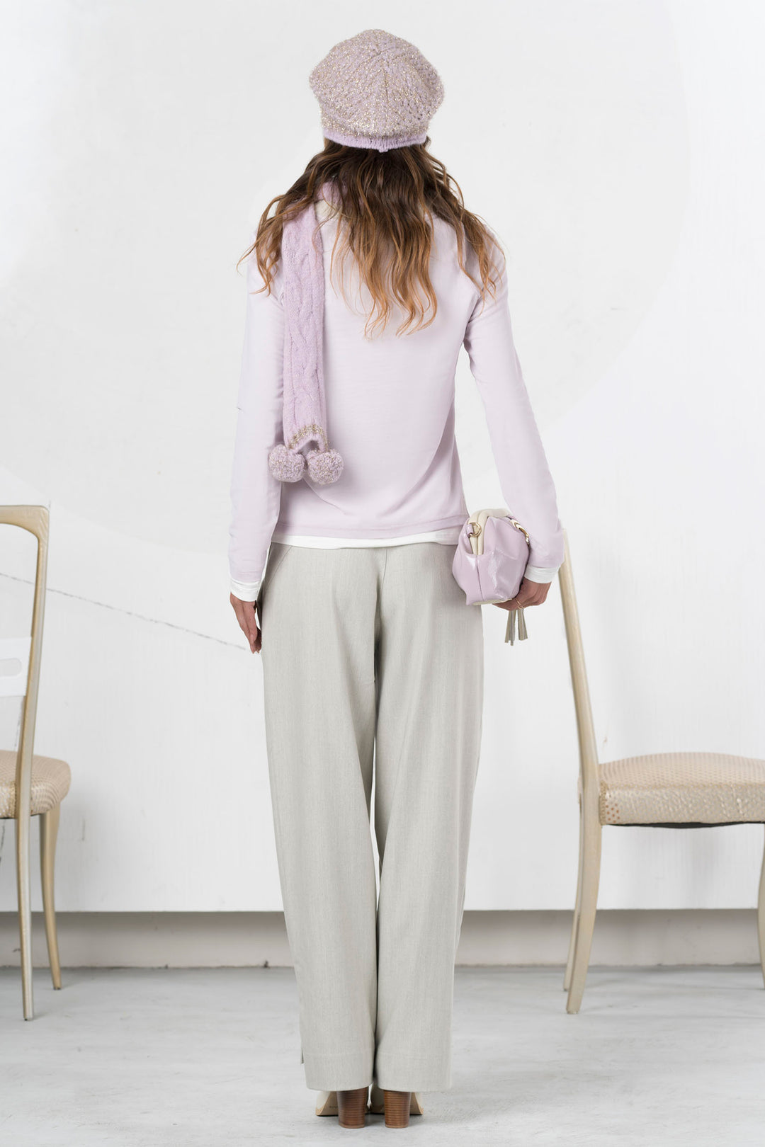 Woman wearing a light pink outfit with a scarf and hat, standing in a minimalistic room.