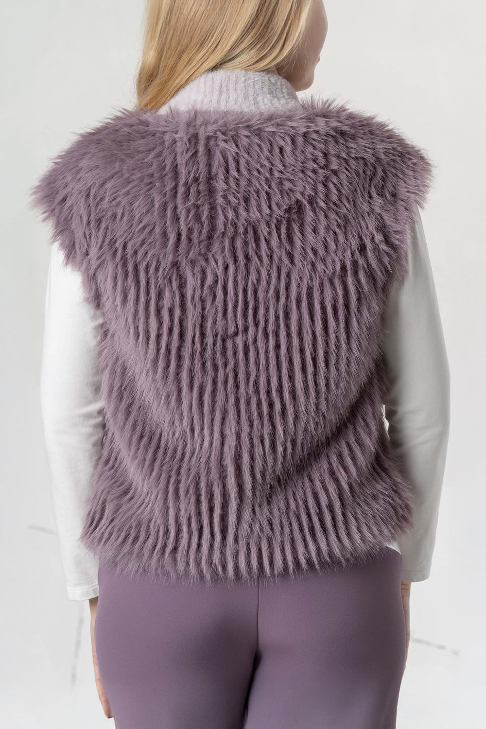 Person wearing a fluffy purple vest over a white shirt and purple pants on a light gray background
