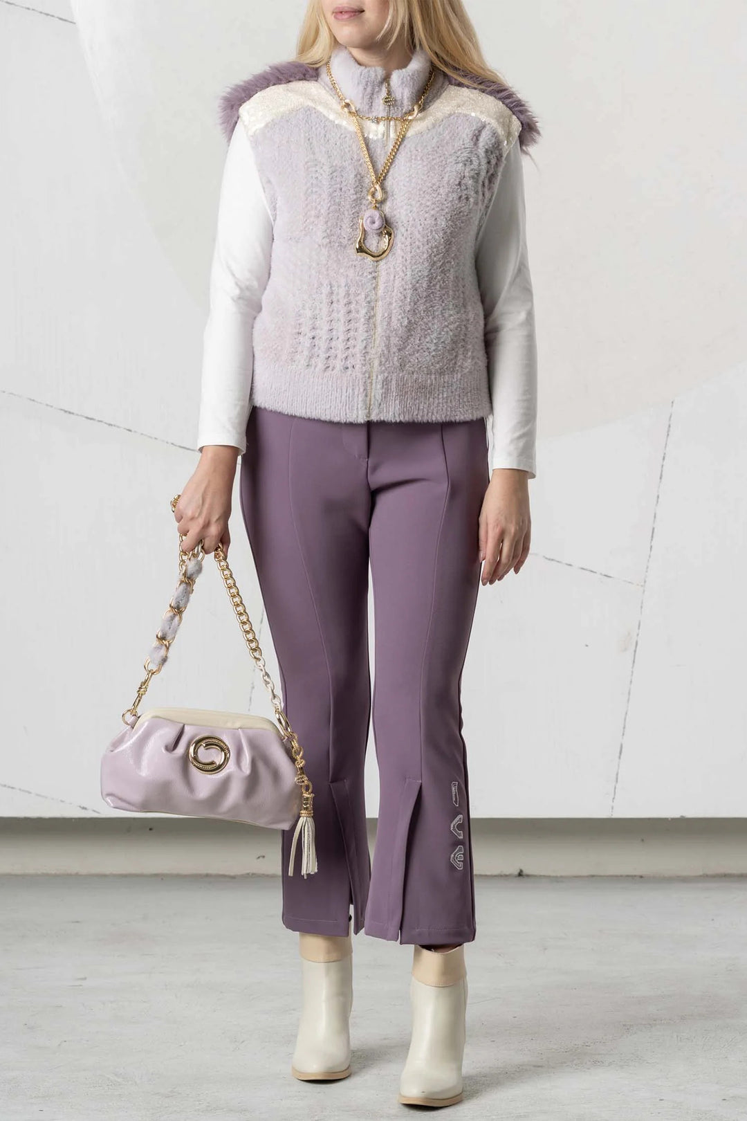 Woman wearing a light purple sweater vest over a white shirt, paired with purple pants and holding a matching handbag.