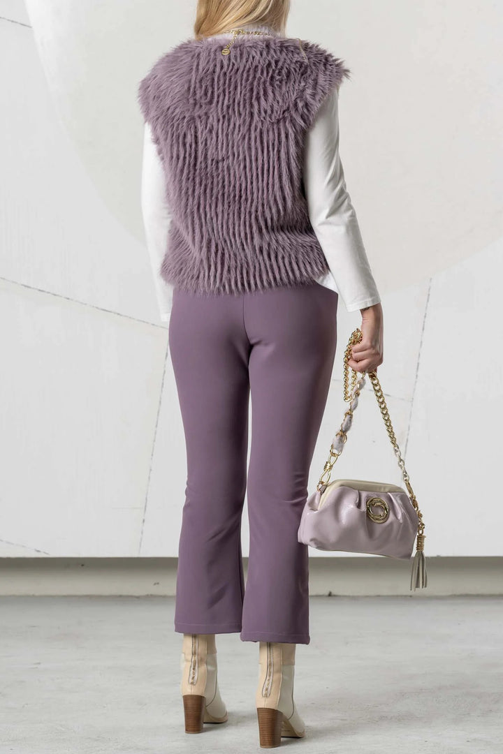 Person wearing a fur vest and purple pants holding a handbag against a white background