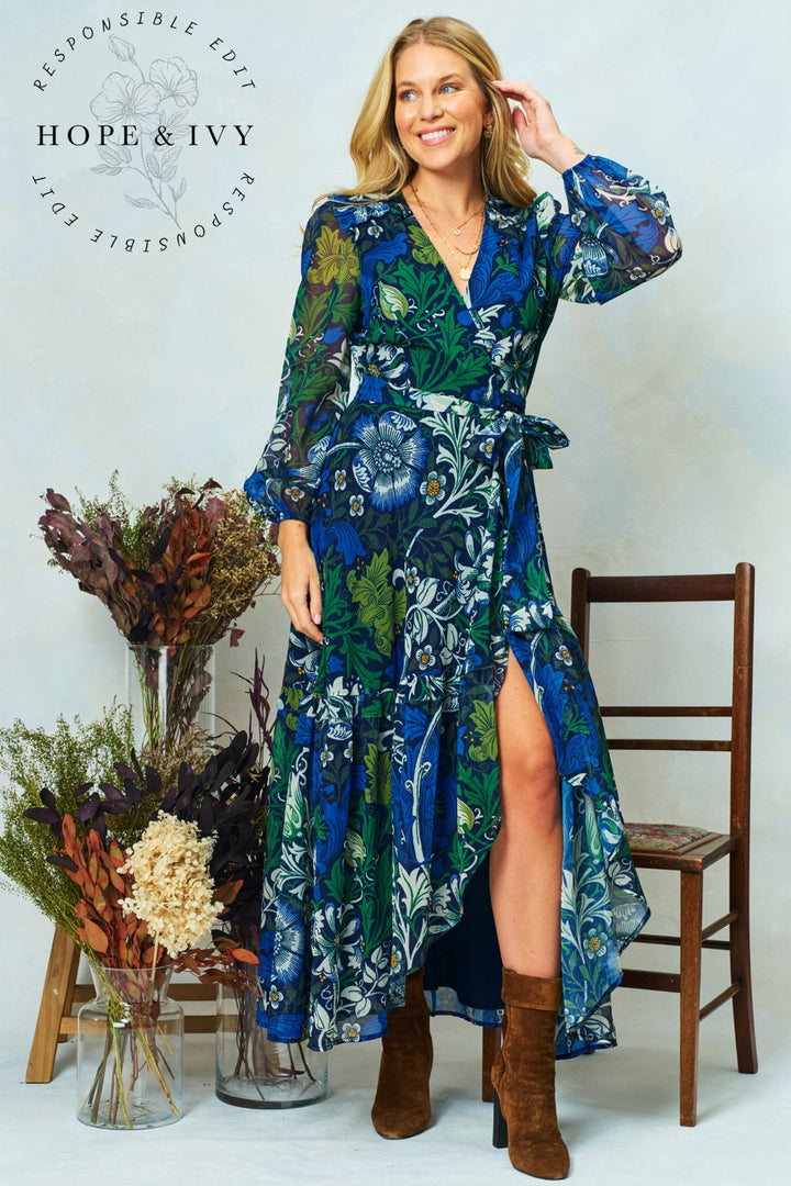 Woman wearing a blue floral dress with brown boots standing next to a wooden chair and dried plants.