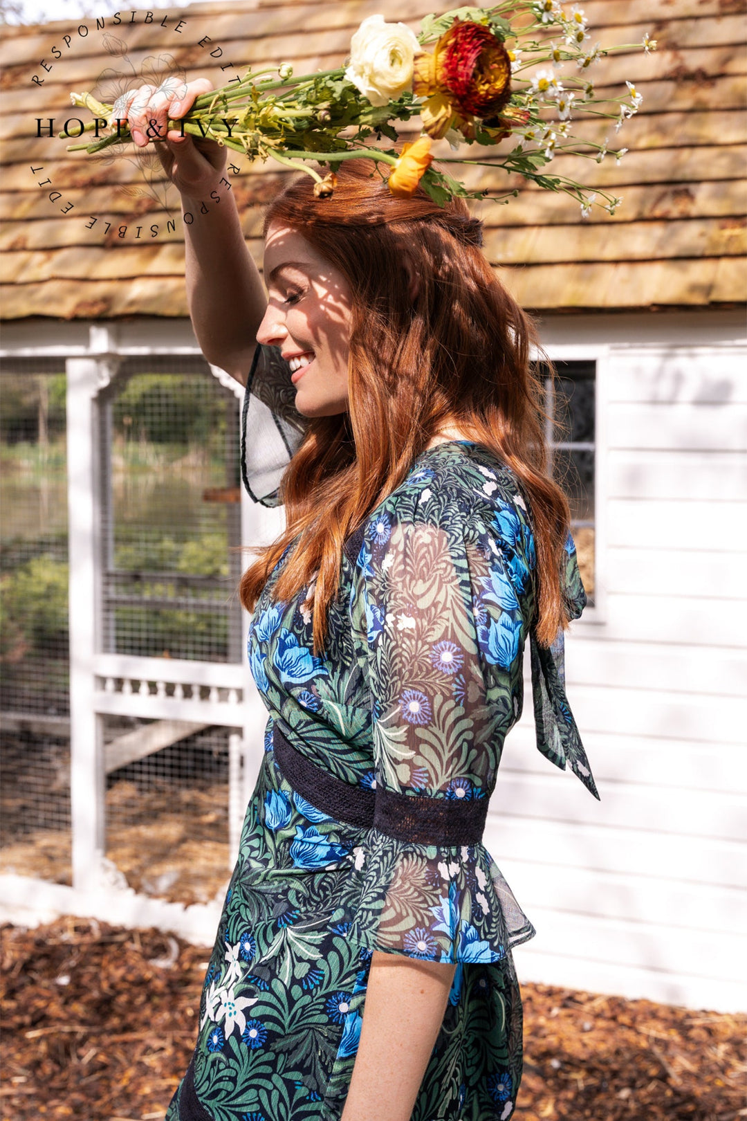 Woman in a floral dress holding flowers outdoors with a rustic background
