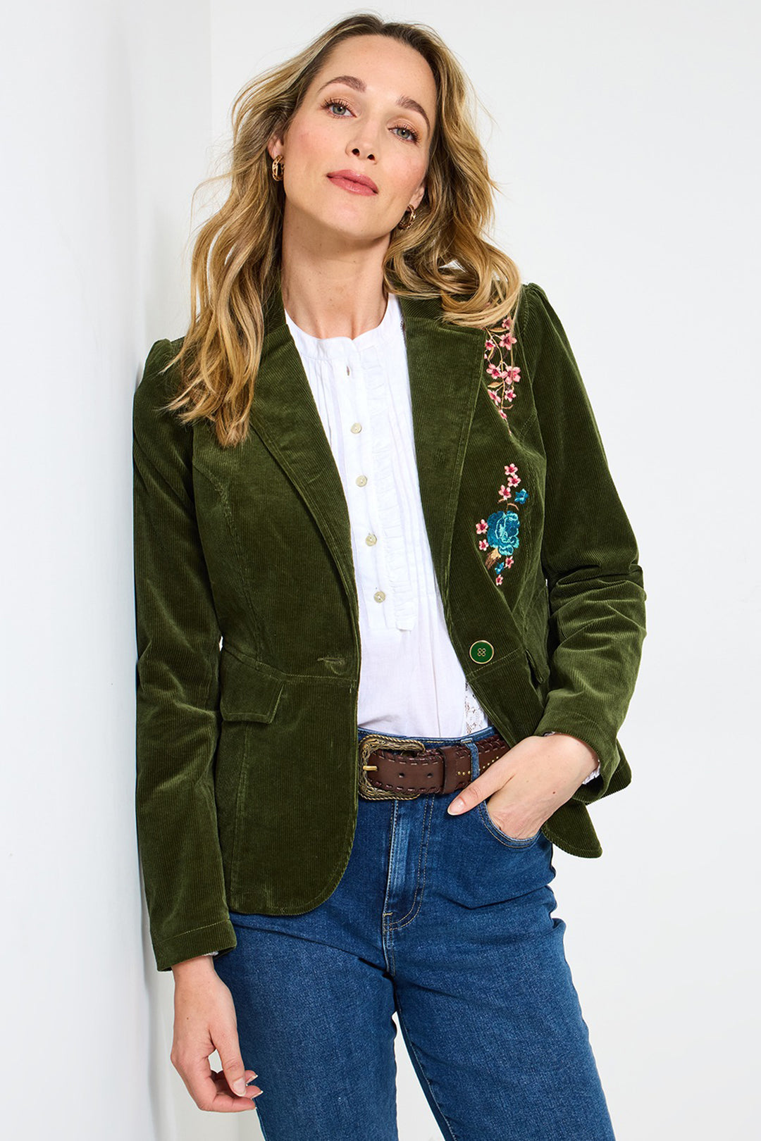 Woman wearing a green blazer with floral embroidery, white shirt, and blue jeans against a white background