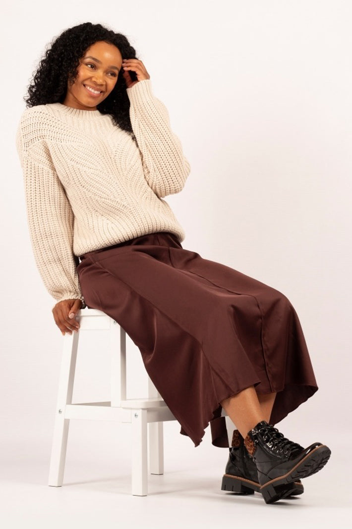Woman sitting on a stool wearing a cream sweater, brown pants, and black boots against a white background
