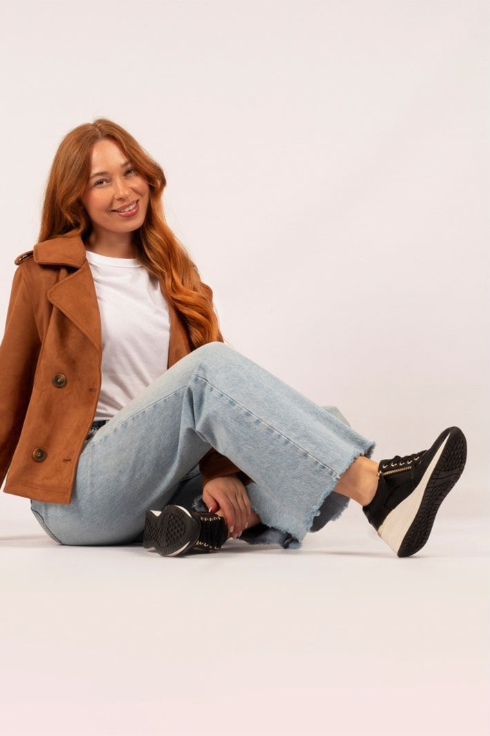 Woman sitting on a white floor wearing a brown coat, white shirt, blue jeans, and black sneakers.