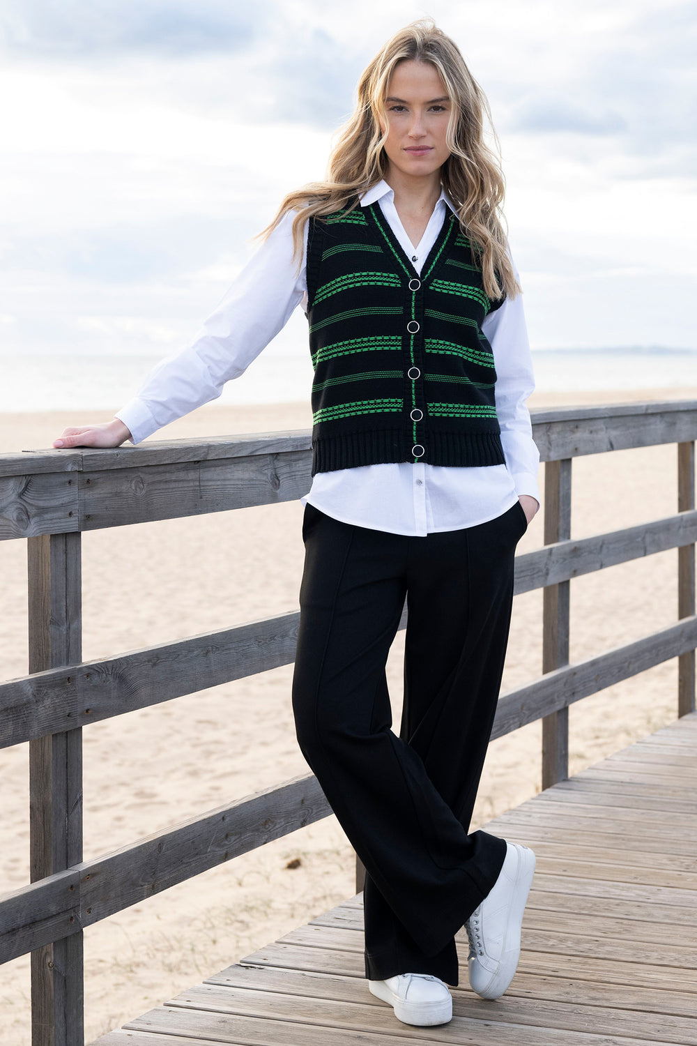 Woman wearing a green patterned vest over a white shirt on a wooden deck by the beach.