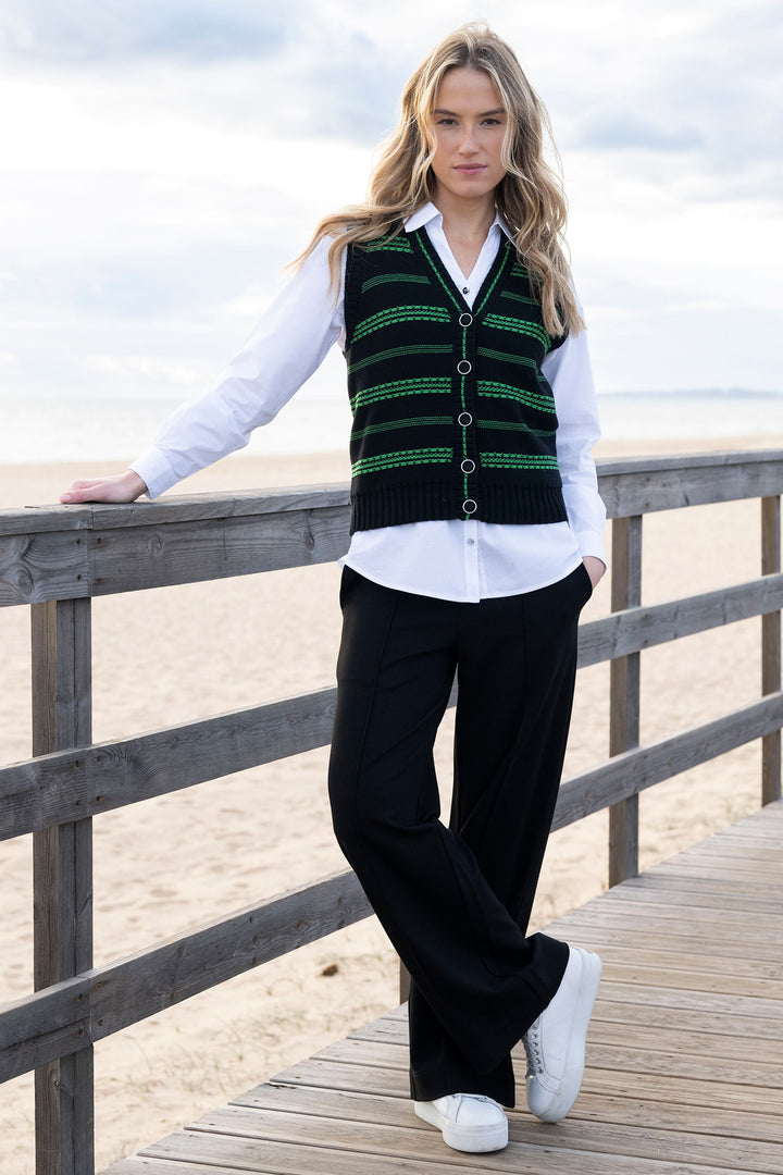 Woman wearing a green patterned vest over a white shirt on a wooden deck by the beach.