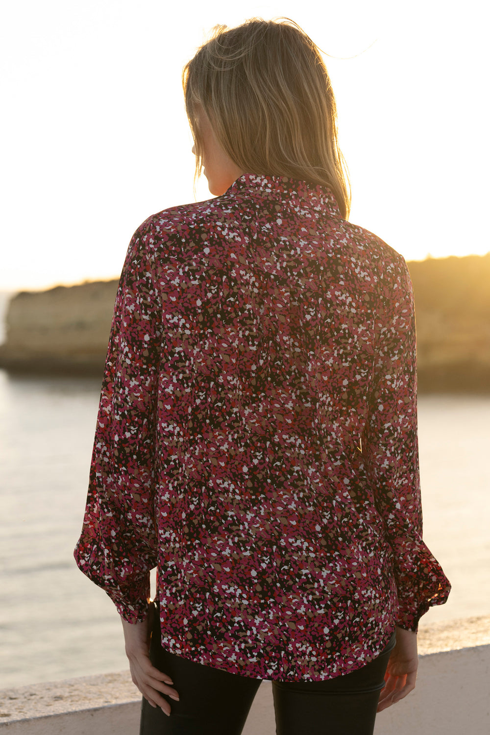Person wearing a floral blouse by a body of water with a sunset.
