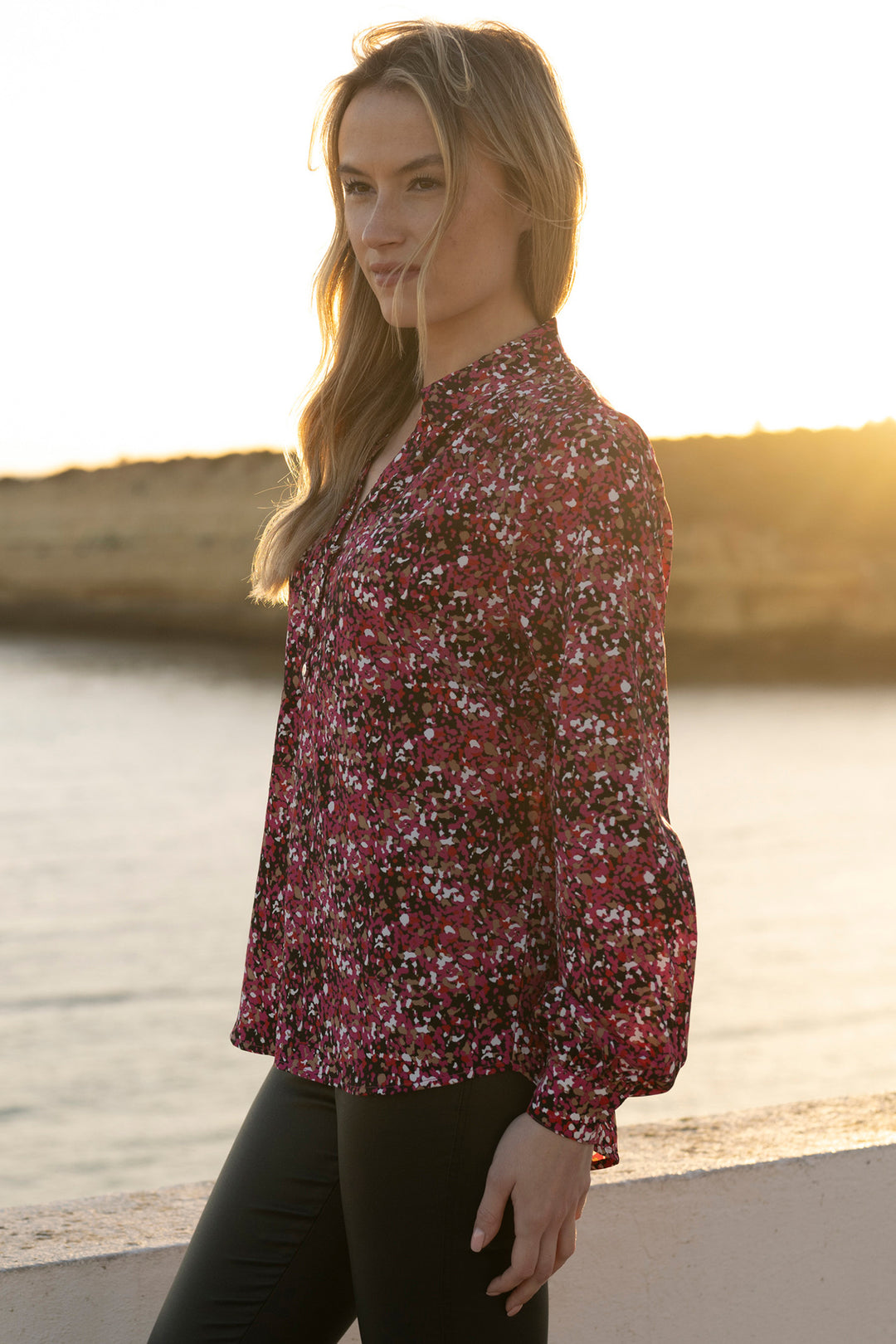 Woman wearing a floral blouse by a body of water with a sunset in the background