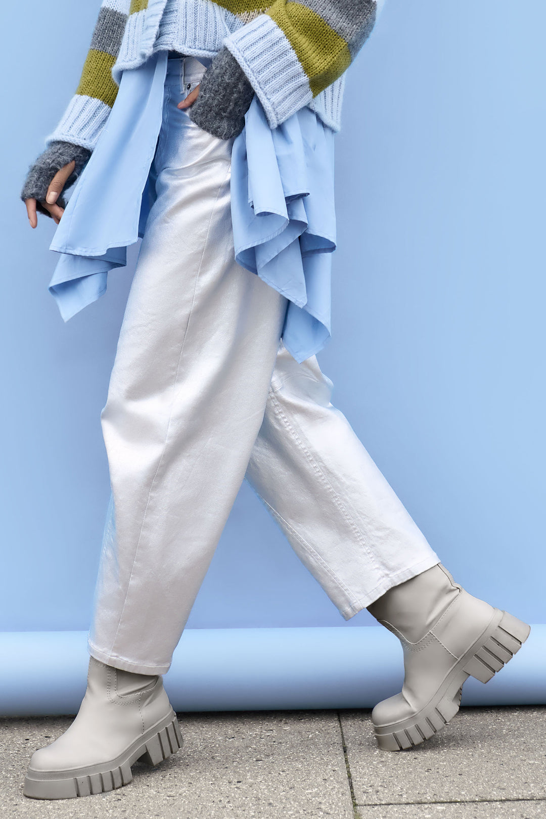 Person wearing silver jeans, grey chunky boots, and a colourful sweater with a blue jacket over the shoulders against a light blue background.