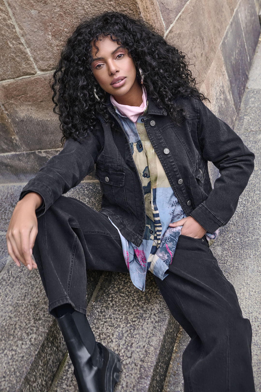 Woman sitting on stone steps wearing a black jacket and pants with a colorful scarf.