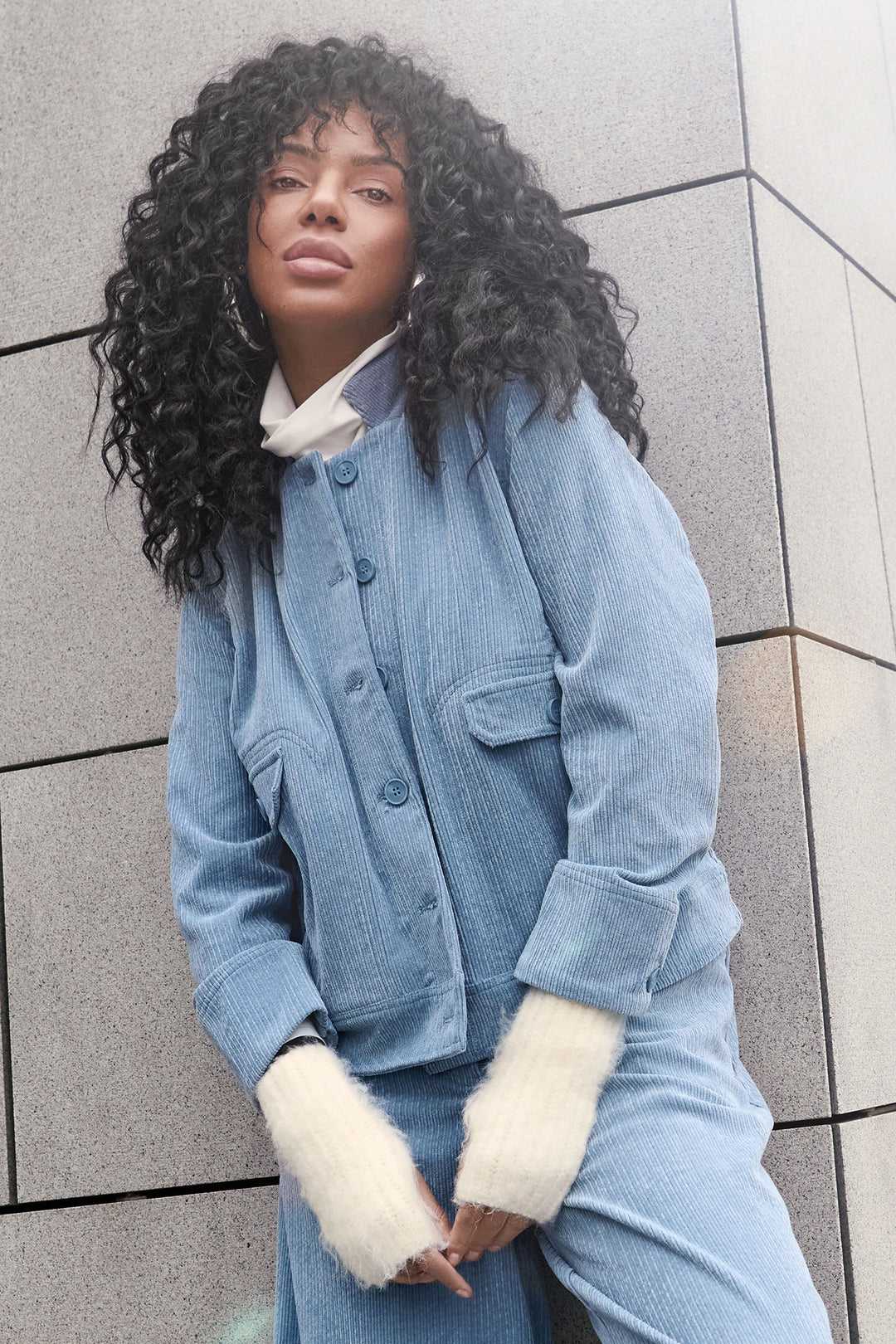 Person wearing a blue denim outfit with white cuffs against a gray tiled wall.