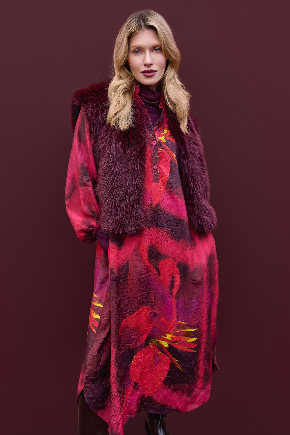 Woman wearing a long, patterned coat with a fur vest against a dark background